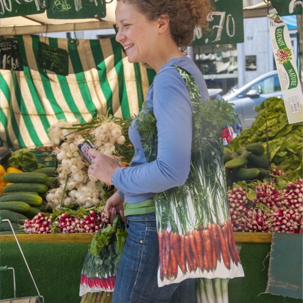 Young woman carrying a Maron Bouillie carrots bag on her shoulder in front of a vegetable stall.