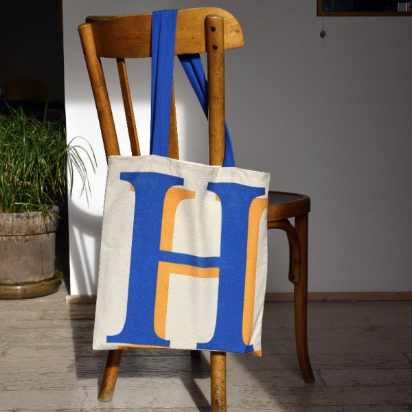 Letter H bag hanging on the back of a wooden chair- French tote bag design by Maron Bouillie