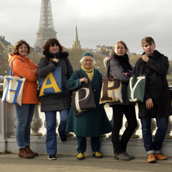 5 people on the street each carrying an initial bag forming the word HAPPY with the Eiffel Tower far away - Maron Bouillie