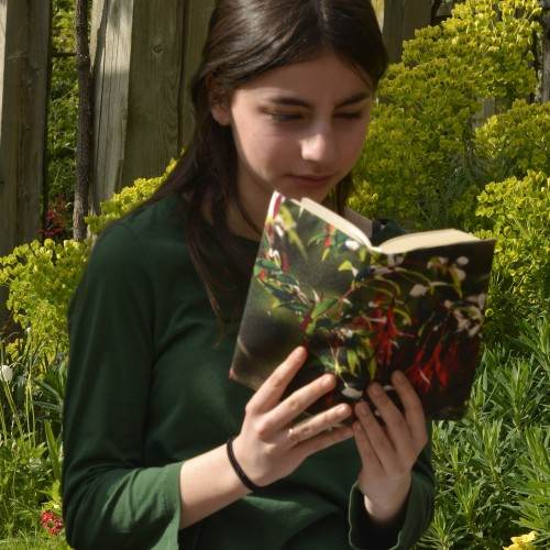 Une jeune fille dans un jardin, lit un livre avec la couverture de livre fleur fuchsia de Maron Bouillie. Plan rapproché
